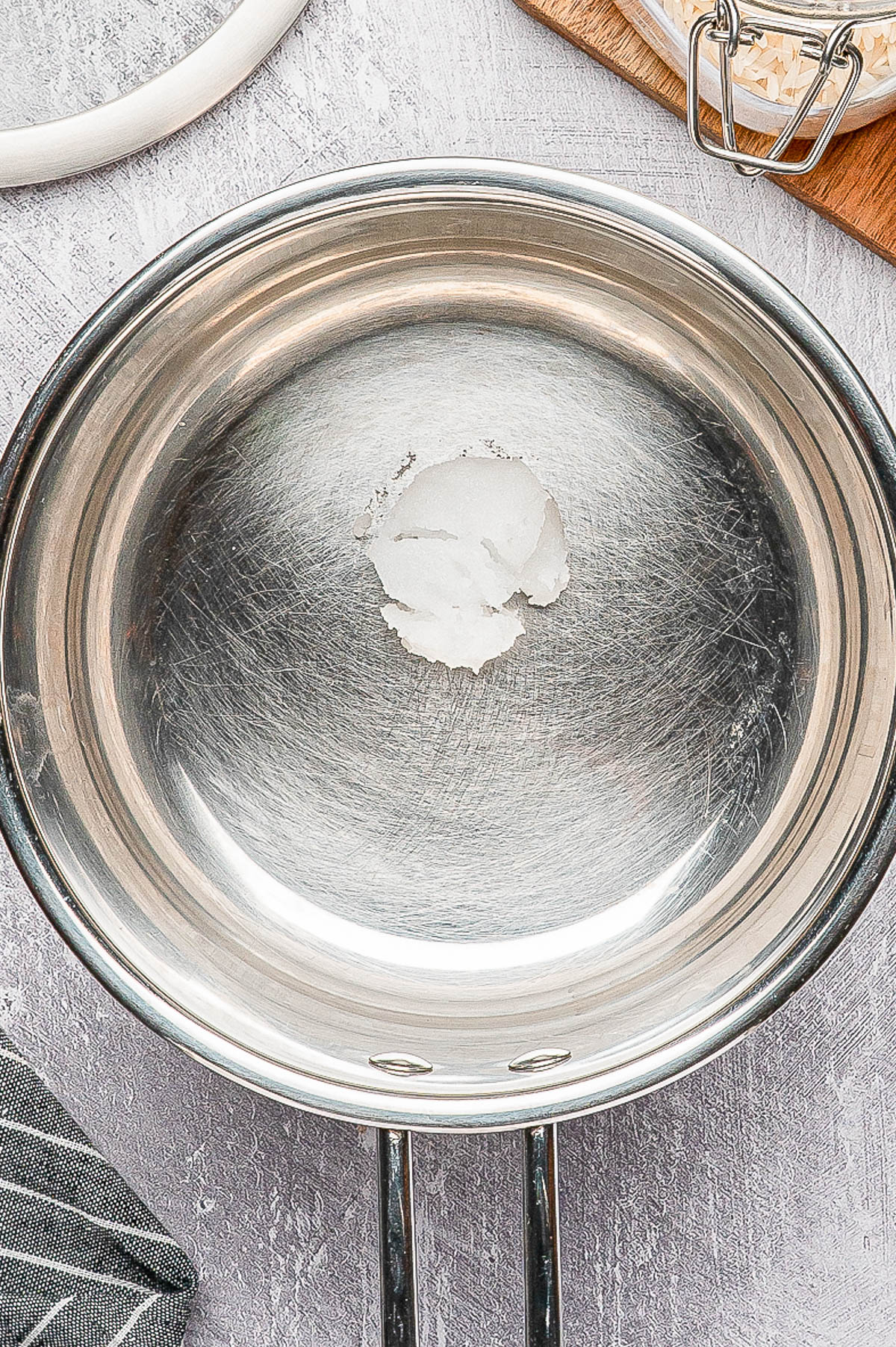 A stainless steel frying pan with a small amount of solidified oil in the center, placed on a gray surface next to a cloth and a wooden board.