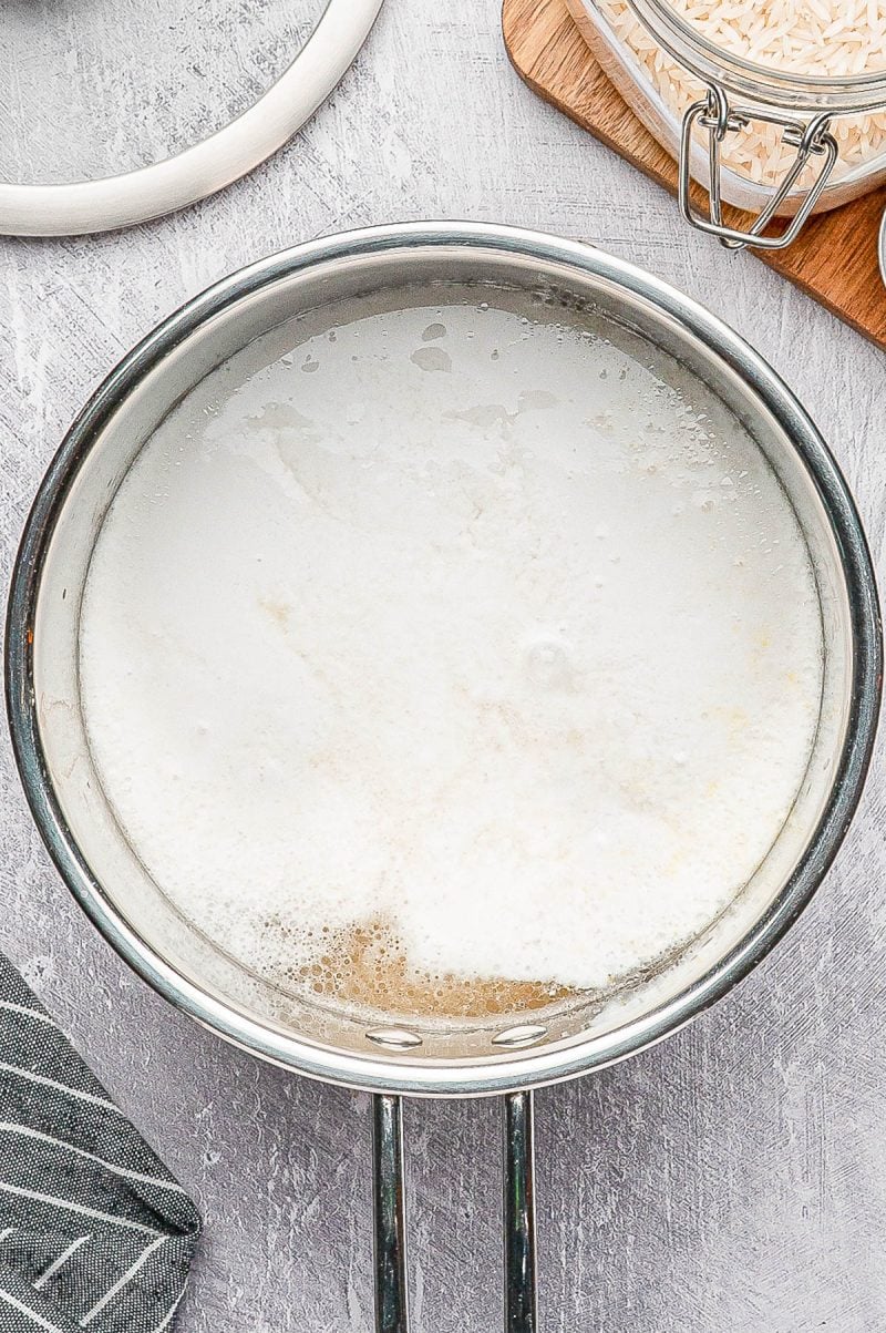 A stainless steel pot filled with boiling water and rice on a gray countertop, with a jar of uncooked rice and a striped cloth nearby.