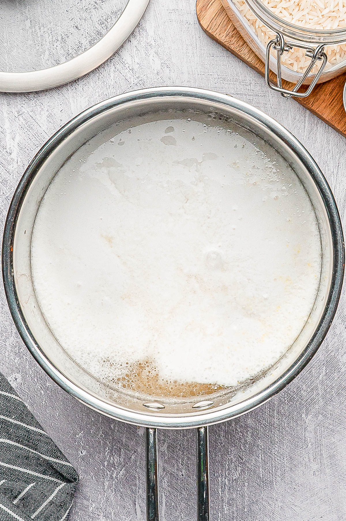 A stainless steel pot filled with boiling water and rice on a gray countertop, with a jar of uncooked rice and a striped cloth nearby.