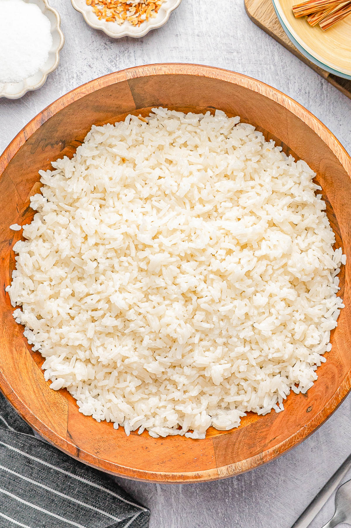 A wooden bowl filled with cooked white rice on a light-colored surface, with a striped napkin and small dishes nearby.