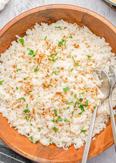 A wooden bowl filled with cooked white rice garnished with chopped herbs and toasted coconut, with two metal serving spoons placed inside.