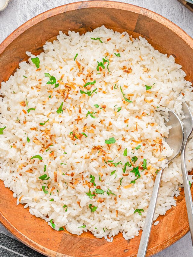 A wooden bowl filled with cooked white rice garnished with chopped herbs and toasted coconut, with two metal serving spoons placed inside.