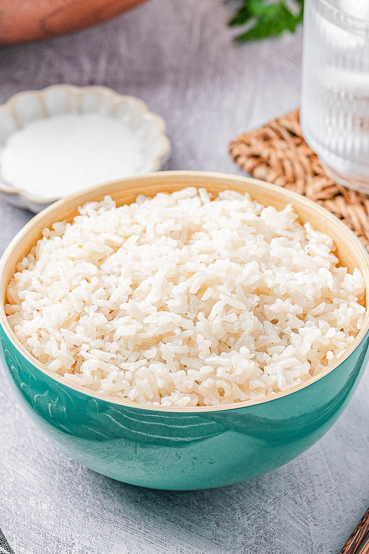 A green bowl filled with cooked white rice sits on a table next to a glass of water and a small dish of salt.