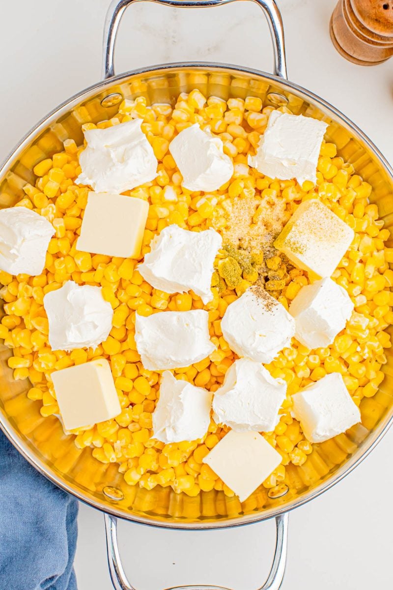 A metal skillet filled with corn kernels, dollops of cream cheese, cubes of butter, and seasonings, ready to be cooked.