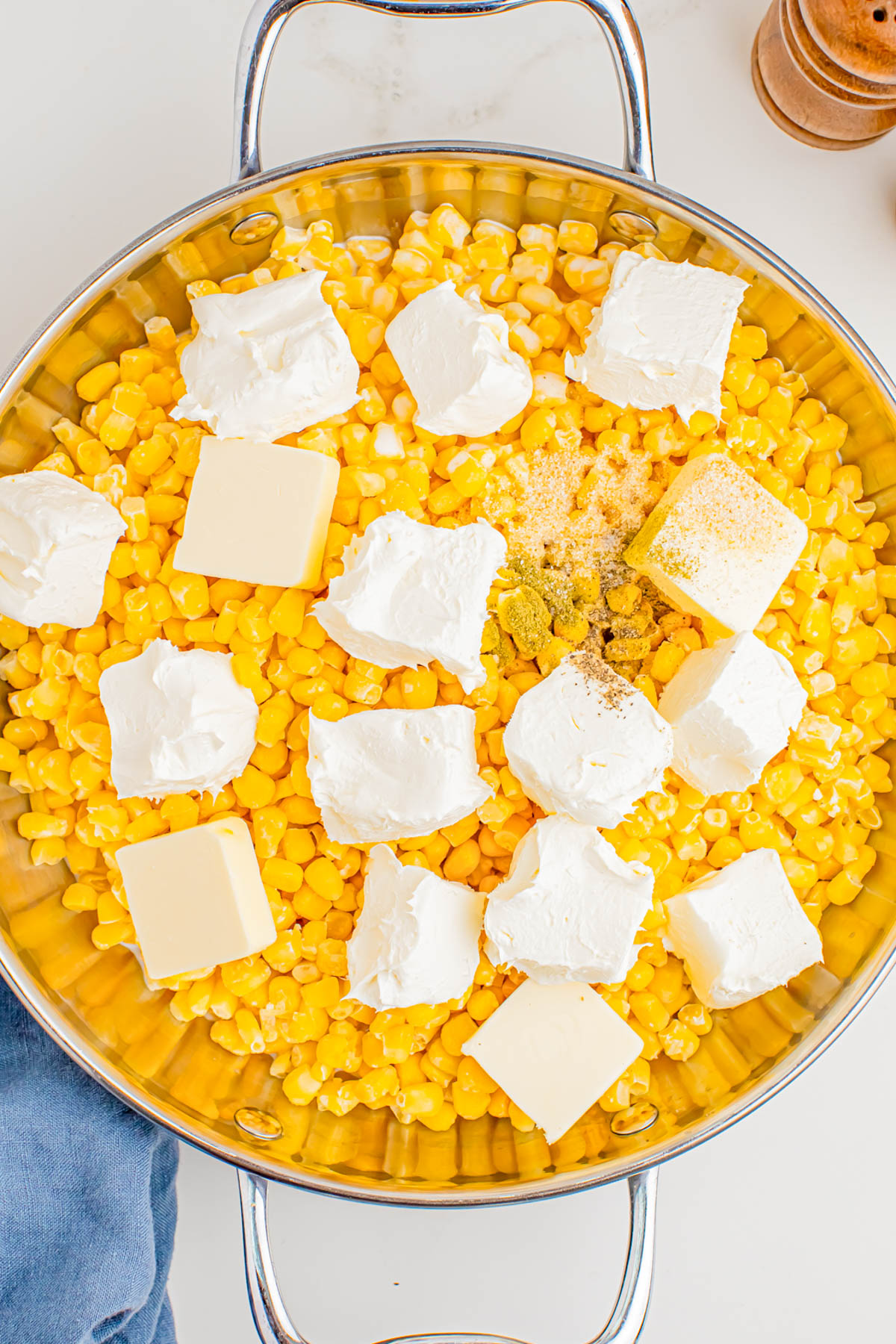 A metal skillet filled with corn kernels, dollops of cream cheese, cubes of butter, and seasonings, ready to be cooked.
