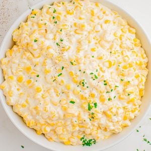 A bowl of creamy corn topped with finely chopped parsley, set on a white surface with a blue napkin and herb sprigs nearby.