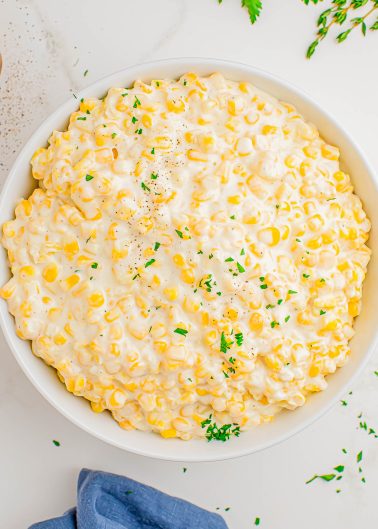 A bowl of creamy corn topped with finely chopped parsley, set on a white surface with a blue napkin and herb sprigs nearby.