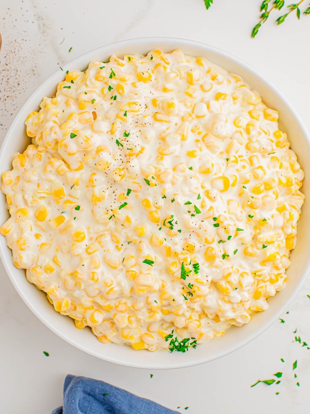 A bowl of creamy corn topped with finely chopped parsley, set on a white surface with a blue napkin and herb sprigs nearby.