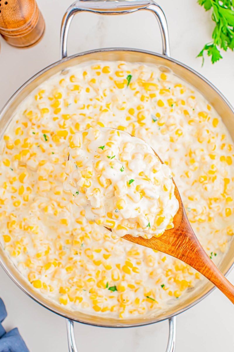 A wooden spoon holds a serving of creamy corn in a large metal pan, garnished with chopped herbs.