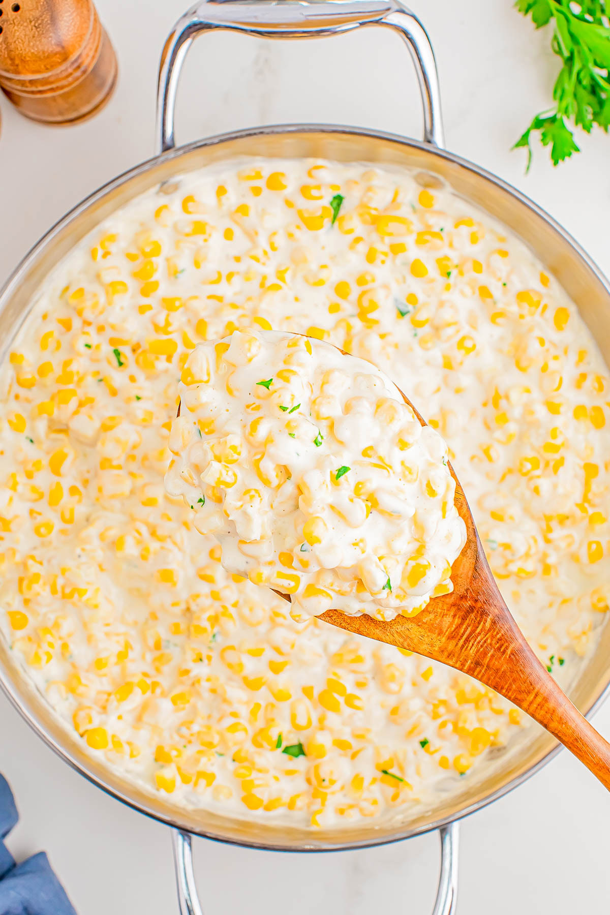 A wooden spoon holds a serving of creamy corn in a large metal pan, garnished with chopped herbs.