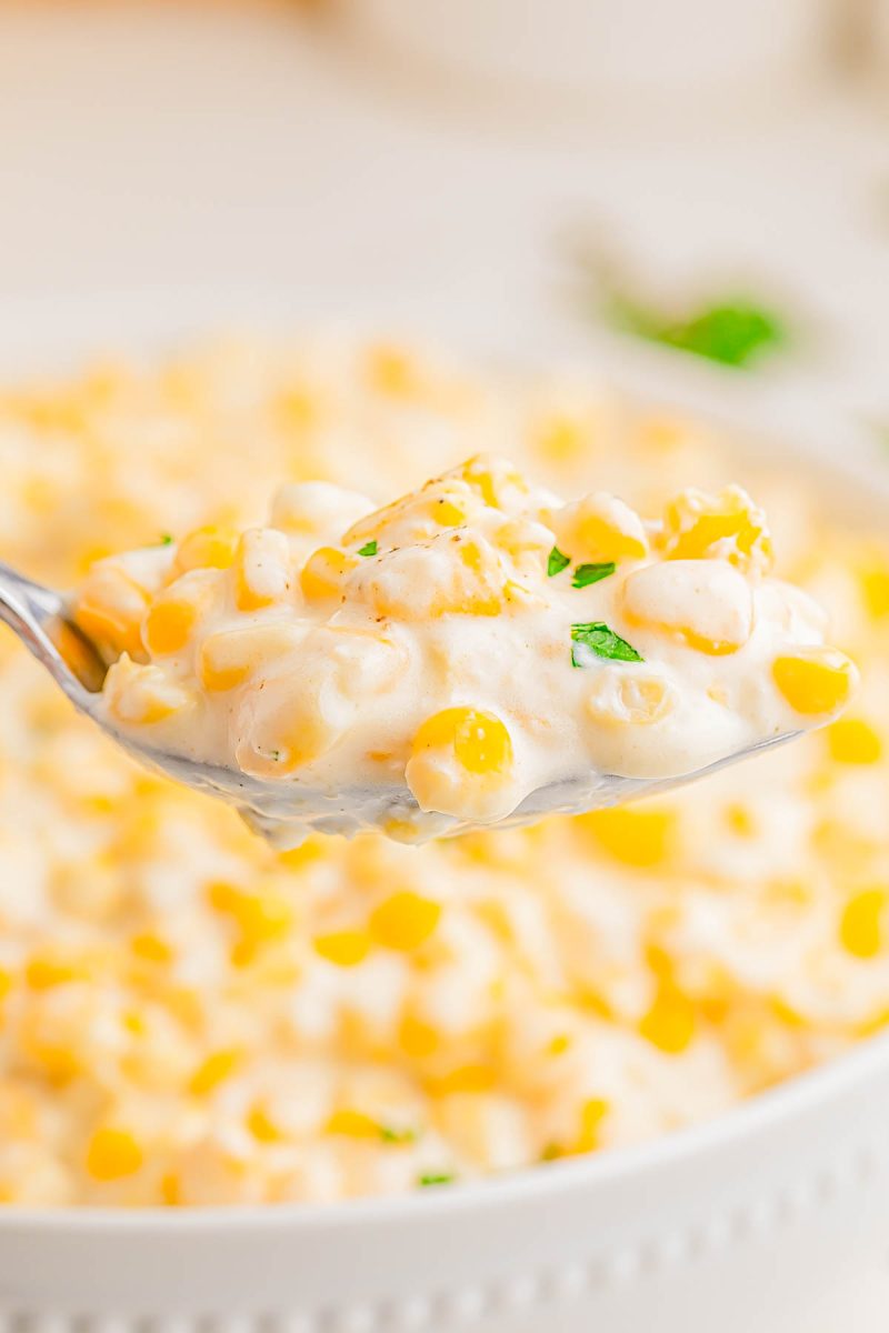 A spoonful of creamy corn casserole garnished with chopped parsley, held above a dish filled with more of the casserole.