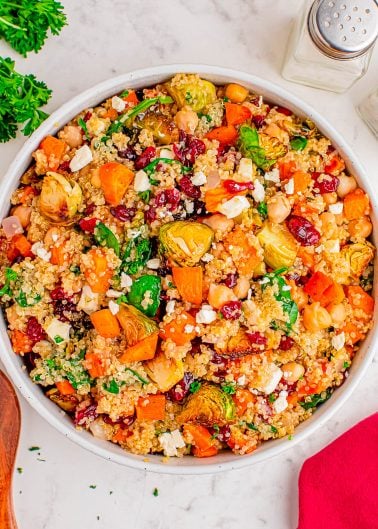 A bowl of quinoa salad with roasted sweet potatoes, Brussels sprouts, chickpeas, dried cranberries, feta cheese, and fresh herbs on a marble surface.