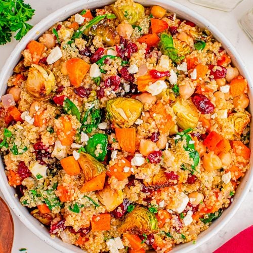 A bowl of quinoa salad with roasted sweet potatoes, Brussels sprouts, chickpeas, dried cranberries, feta cheese, and fresh herbs on a marble surface.