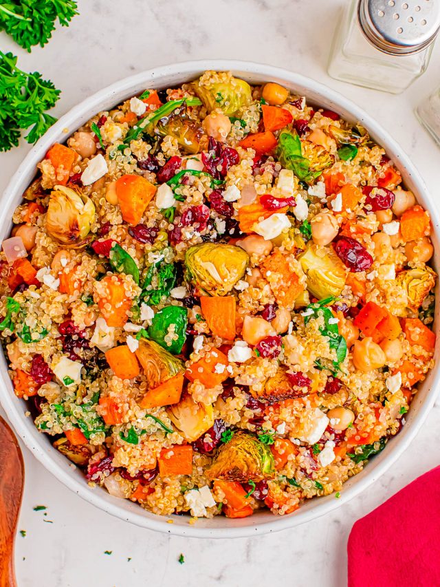 A bowl of quinoa salad with roasted sweet potatoes, Brussels sprouts, chickpeas, dried cranberries, feta cheese, and fresh herbs on a marble surface.