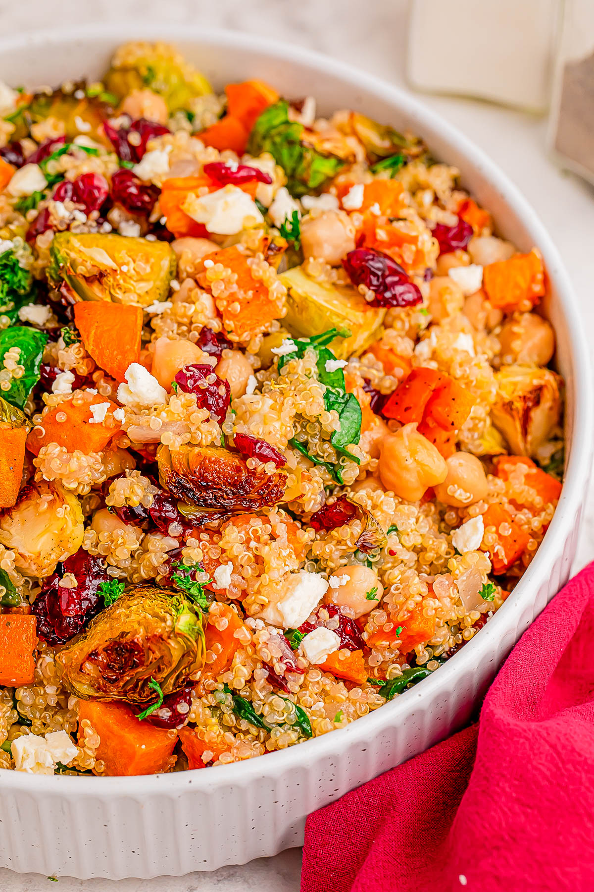 A white dish filled with quinoa salad, featuring roasted Brussels sprouts, sweet potatoes, chickpeas, dried cranberries, chopped greens, and crumbled feta cheese.
