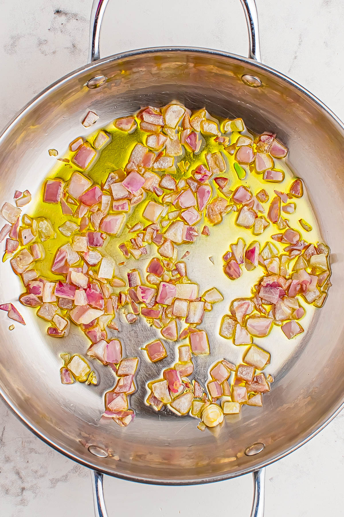 Chopped red onions sautรฉing in olive oil in a stainless steel pan on a light-colored surface.