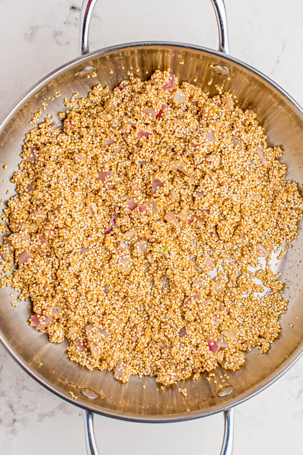 A stainless steel pan containing uncooked quinoa mixed with diced onions is shown on a light countertop.