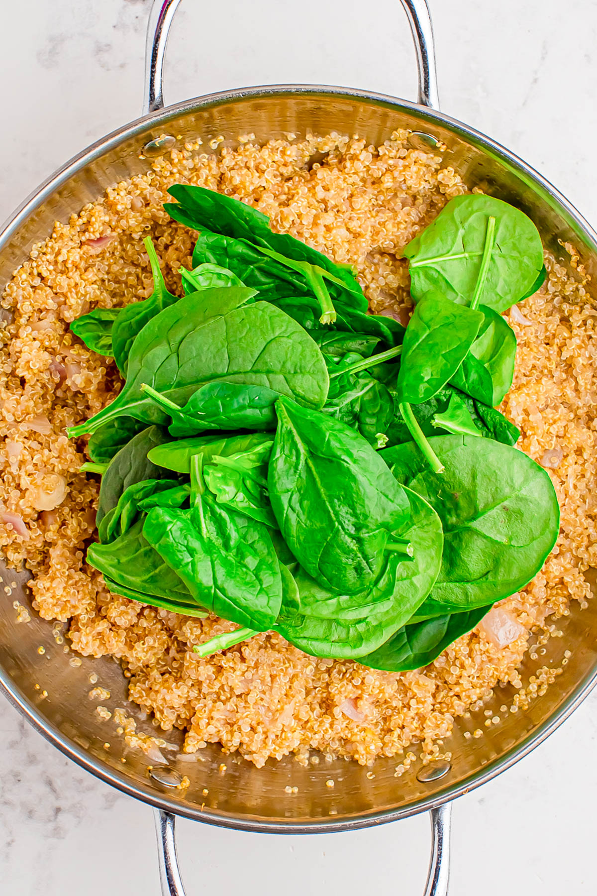 A stainless steel pan containing cooked quinoa with raw spinach leaves on top, ready to be mixed in.