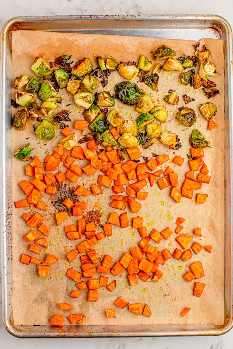 Roasted Brussels sprouts and diced sweet potatoes are spread on a parchment-lined baking sheet.