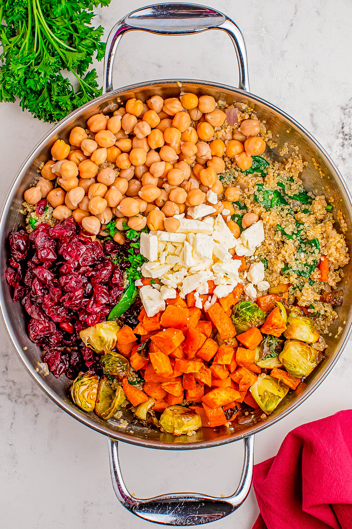 A skillet containing chickpeas, dried cranberries, feta cheese, roasted sweet potatoes, Brussels sprouts, and quinoa, arranged in separate sections.