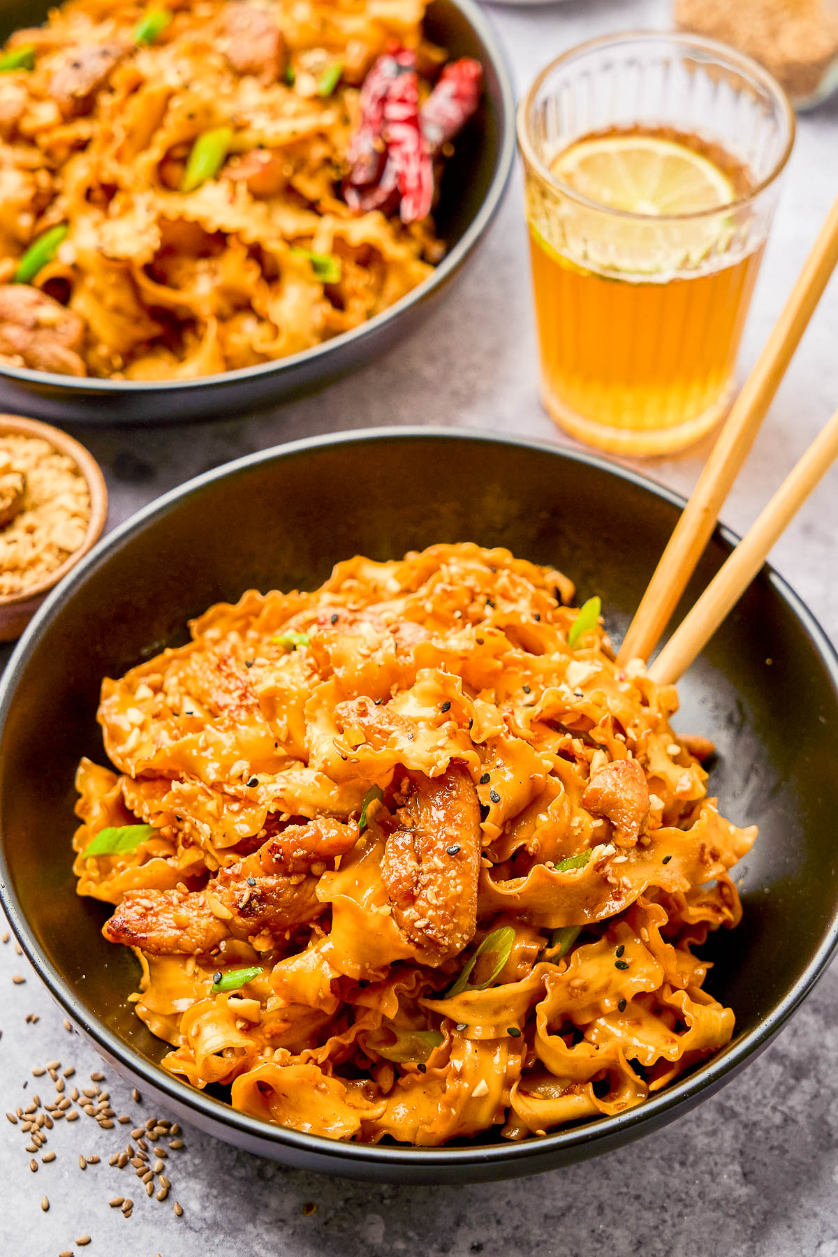 A black bowl filled with wide, ribbon-shaped noodles mixed with sliced chicken and green onions, served with chopsticks; a glass of iced tea and another bowl in the background.