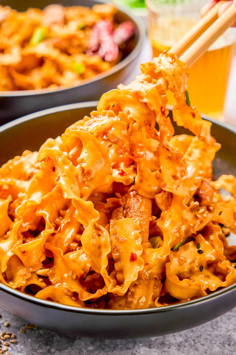A bowl of wide, ruffled noodles coated in a creamy, spicy sauce, with pieces of meat and toppings, being lifted by chopsticks.