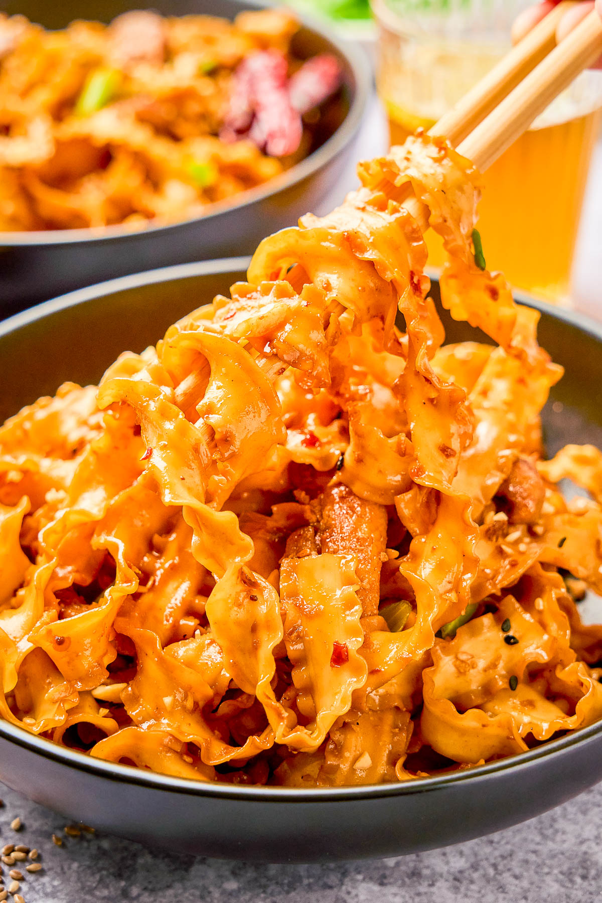 A bowl of wide, ruffled noodles coated in a creamy, spicy sauce, with pieces of meat and toppings, being lifted by chopsticks.