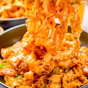 A bowl of spicy, saucy noodles with pieces of meat and green onions, being lifted by chopsticks.