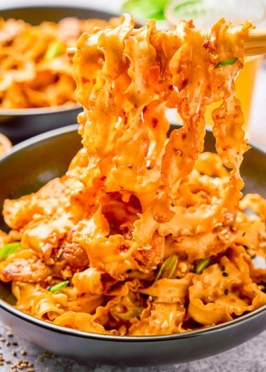 A bowl of spicy, saucy noodles with pieces of meat and green onions, being lifted by chopsticks.