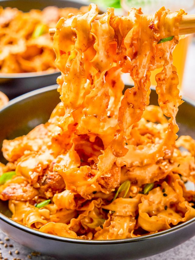 A bowl of spicy, saucy noodles with pieces of meat and green onions, being lifted by chopsticks.