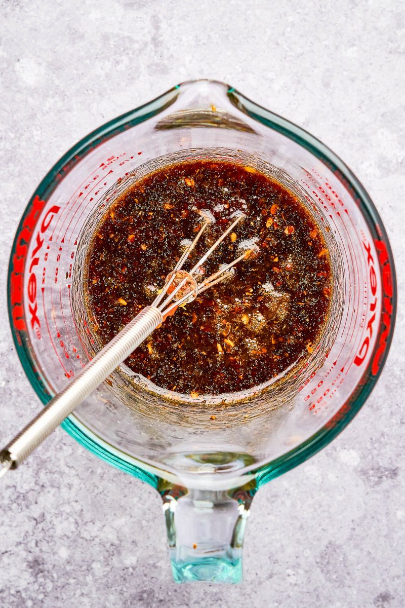 A glass measuring cup filled with a dark, mixed liquid sauce and a metal whisk inside on a light gray surface.