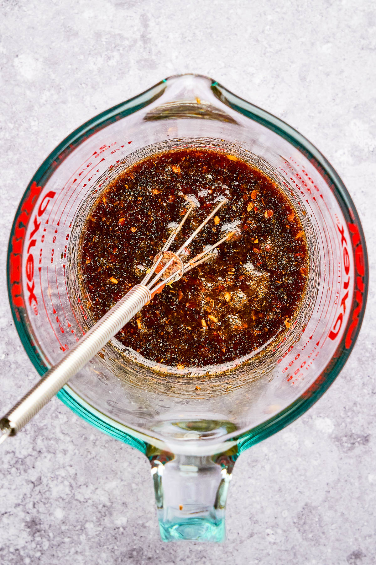 A glass measuring cup filled with a dark, mixed liquid sauce and a metal whisk inside on a light gray surface.