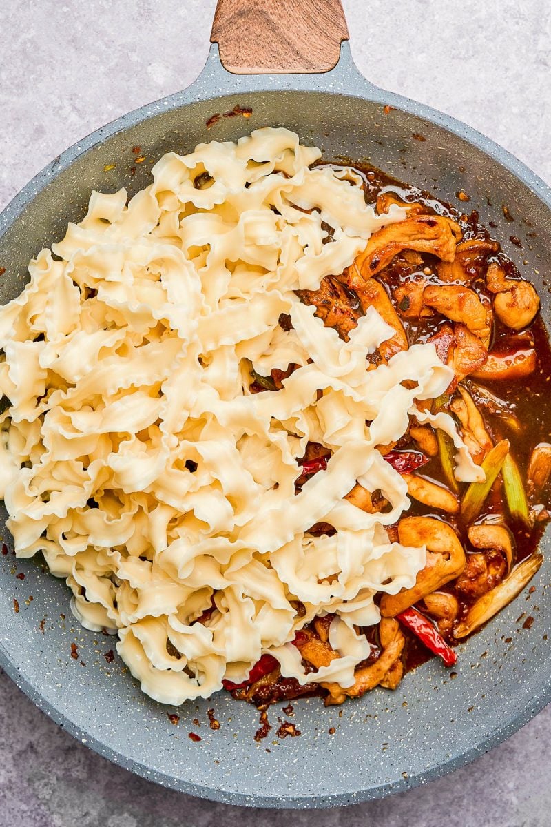 A skillet with uncooked wide pasta on one side and cooked shrimp with vegetables and sauce on the other side.