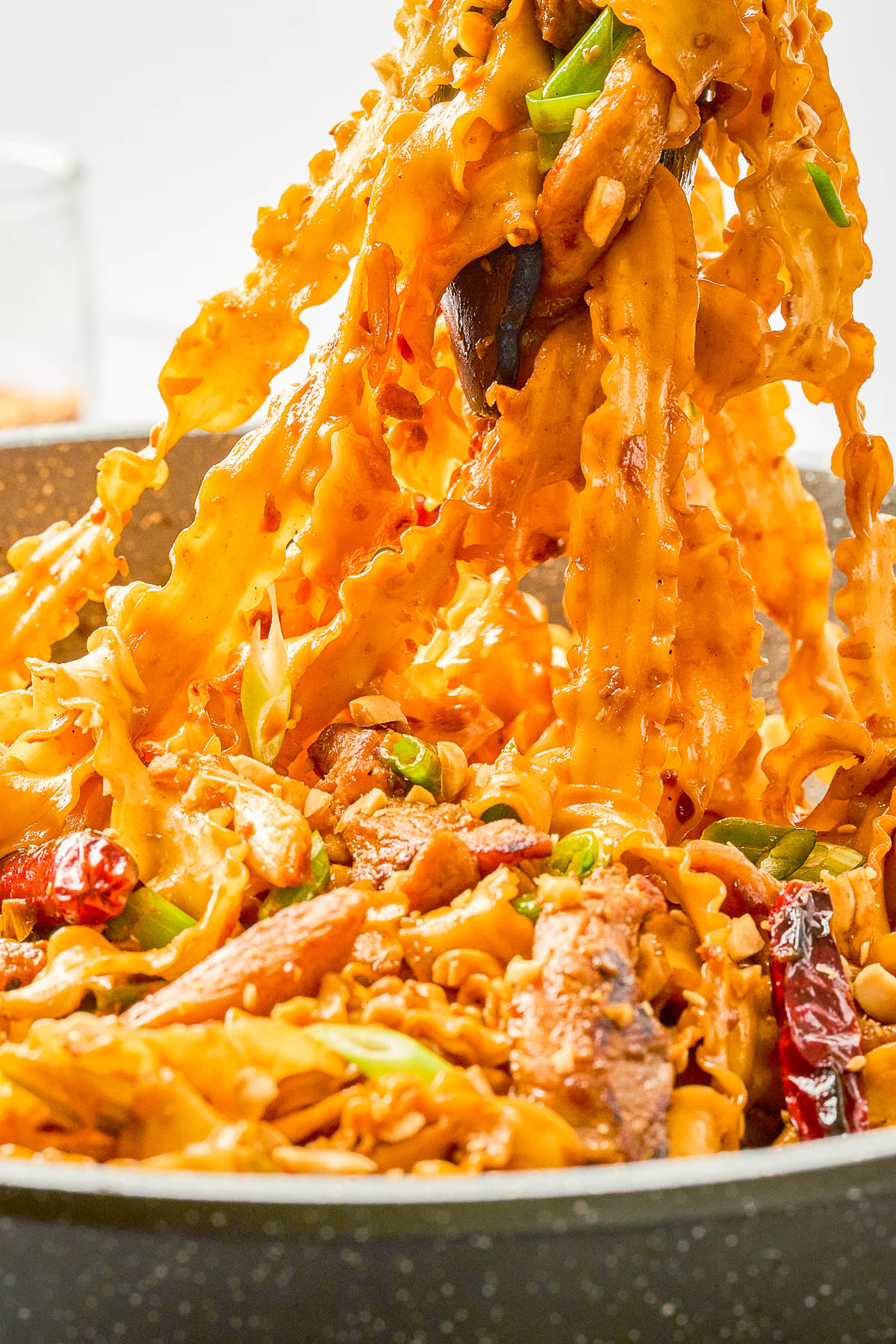 Wide, curly pasta noodles coated in a creamy orange sauce with pieces of meat, dried chili peppers, and green onions, being lifted from a pan.