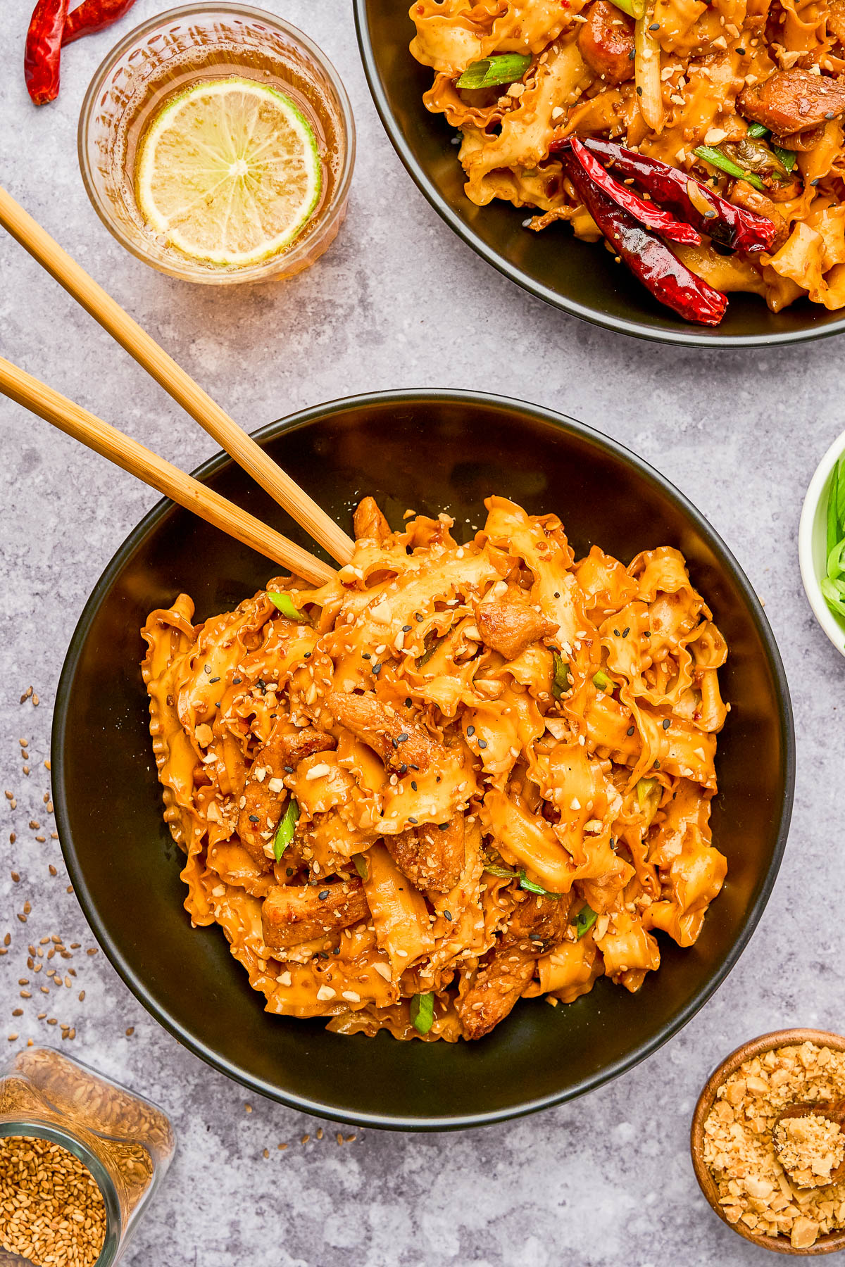 A bowl of spicy noodles with pieces of chicken, garnished with sesame seeds and green onions, with wooden chopsticks resting on top. Nearby are a drink, a bowl of noodles, and condiments.
