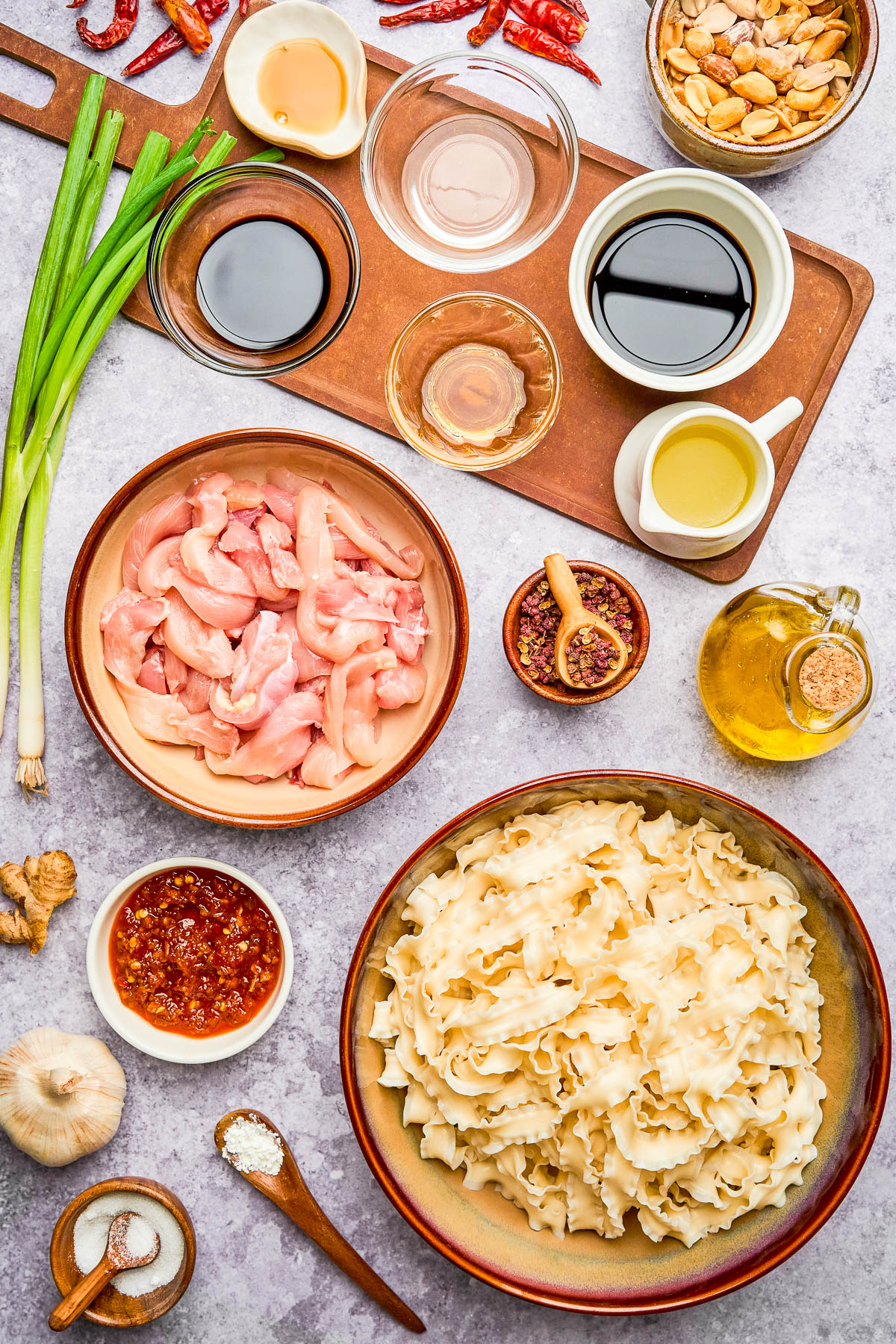 Ingredients for a noodle dish arranged on a table, including raw chicken, wide noodles, sauces, oil, garlic, ginger, scallions, peanuts, and spices.