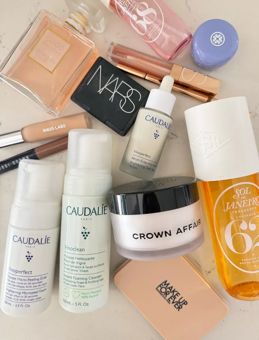 A flat lay of various skincare and makeup products, including Caudalie, Sol de Janeiro, NARS, MAKE UP FOR EVER, Haus Labs, and others, arranged on a light surface.