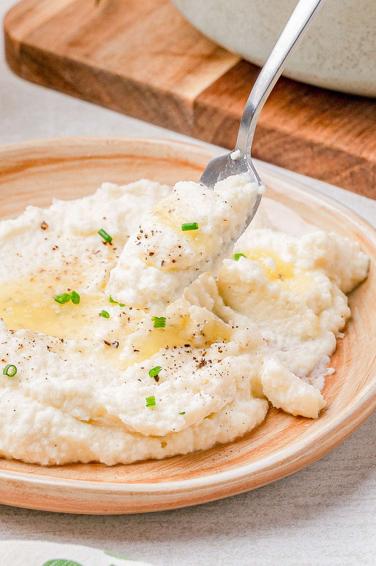 A plate of mashed cauliflower topped with melted butter, chopped chives, and ground black pepper; a fork lifts a bite from the plate.