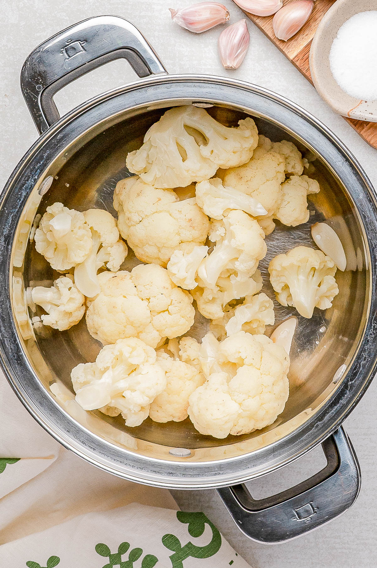A stainless steel pot filled with cauliflower florets, placed on a light surface with garlic cloves and a bowl of salt nearby.