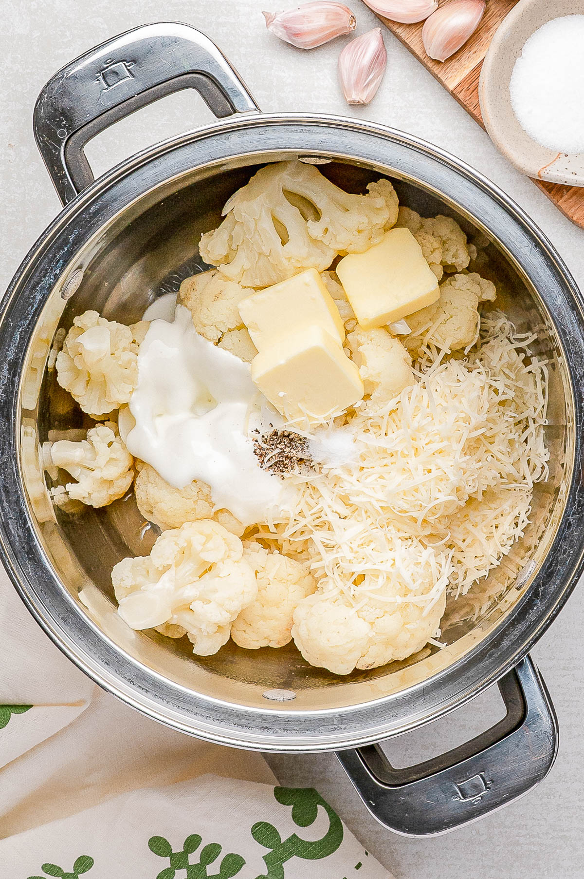 A metal bowl contains cauliflower florets, cubes of butter, shredded cheese, a dollop of sour cream, and ground black pepper, ready to be mixed. Garlic, salt, and a cloth are nearby.