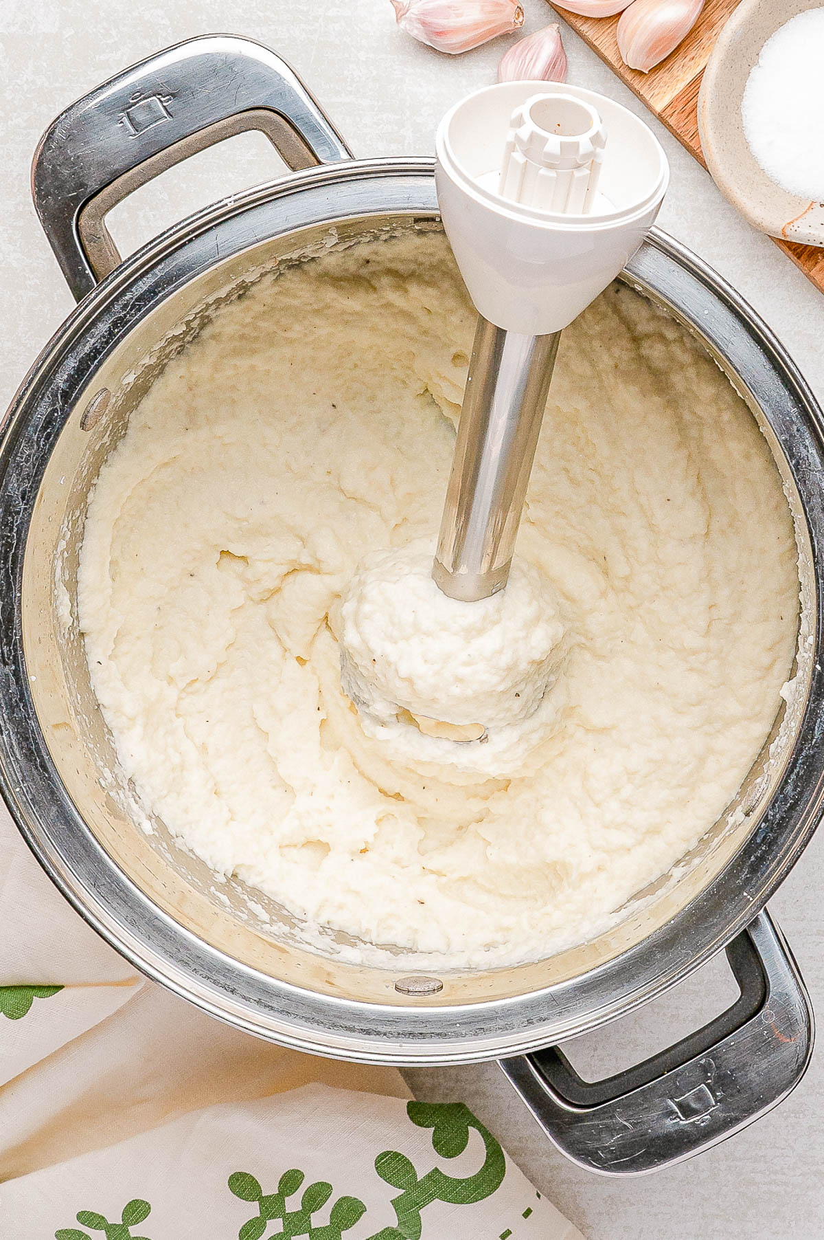An immersion blender is blending mashed potatoes in a large stainless steel pot, with garlic cloves and a bowl of salt nearby.