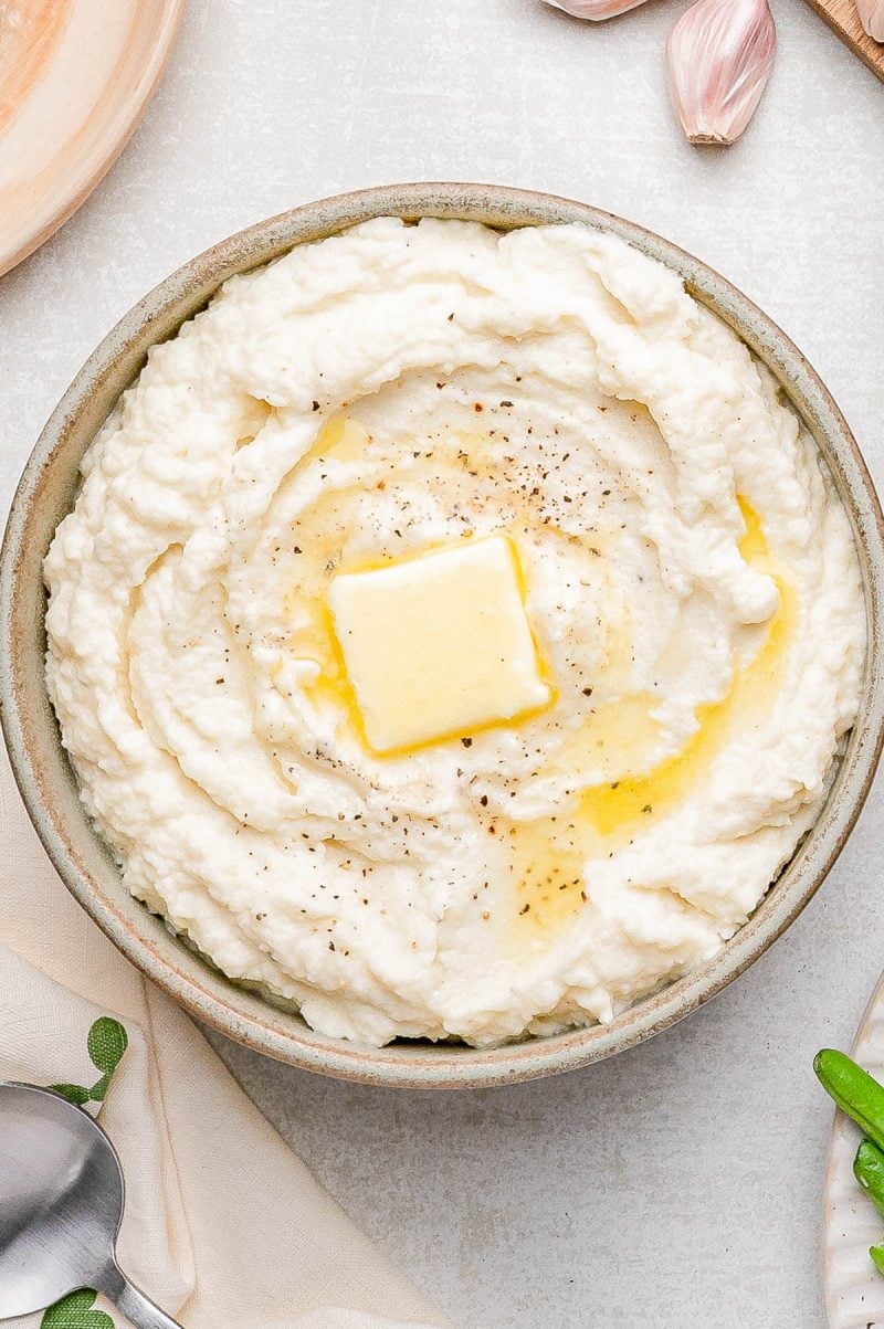A bowl of mashed potatoes topped with a pat of butter and ground black pepper, surrounded by garlic cloves and a spoon on a light surface.
