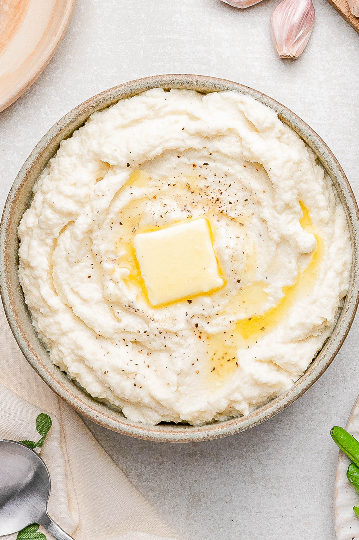 A bowl of mashed potatoes topped with a pat of butter and ground black pepper, surrounded by garlic cloves and a spoon on a light surface.