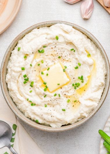 A bowl of mashed potatoes topped with a pat of melting butter, chopped chives, and cracked black pepper.