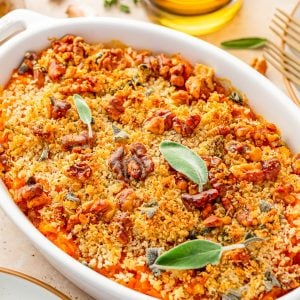 A baked casserole topped with golden breadcrumbs, chopped nuts, and fresh sage leaves, served in a white oval dish on a table.