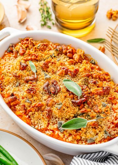A baked casserole topped with golden breadcrumbs, chopped nuts, and fresh sage leaves, served in a white oval dish on a table.