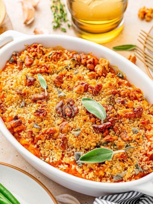 A baked casserole topped with golden breadcrumbs, chopped nuts, and fresh sage leaves, served in a white oval dish on a table.