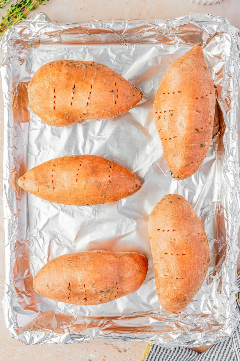 Four whole sweet potatoes with fork holes are placed on a foil-lined baking sheet, ready for roasting.