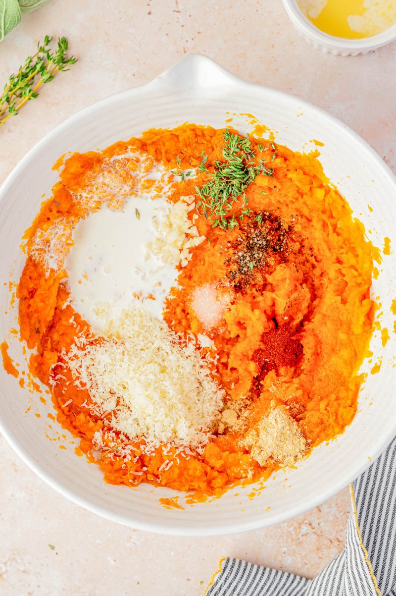 A bowl of mashed sweet potatoes with added cream, grated cheese, spices, minced garlic, salt, pepper, and fresh thyme on top, ready to be mixed.