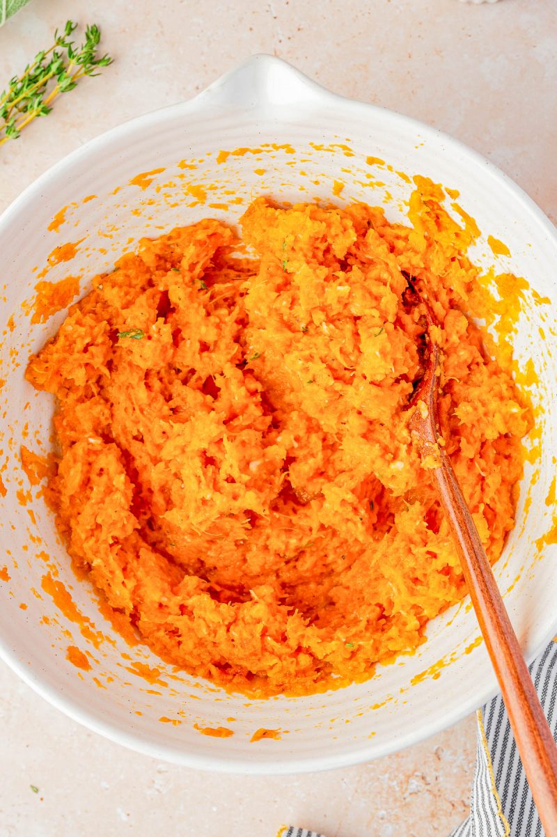 A white mixing bowl filled with a mashed orange mixture being stirred with a wooden spoon.